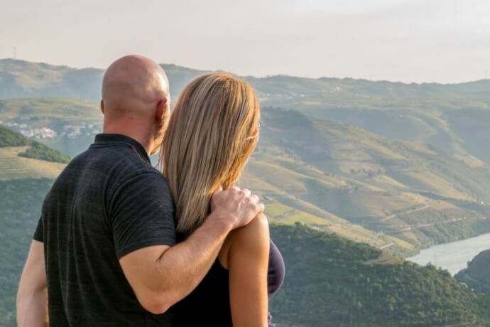 A couple embracing while gazing over the rolling hills and vineyards of Portugal's Douro Valley, with a river winding through the landscape below.