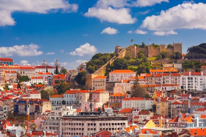 Panoramic view of Lisbon’s Alfama district with São Jorge Castle rising above red-tiled rooftops under a blue sky.