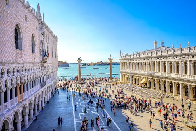 Bright daytime scene of Venice’s bustling Piazzetta San Marco filled with visitors, flanked by the Doge’s Palace and Libreria Sansoviniana, with columns and the sparkling lagoon beyond.