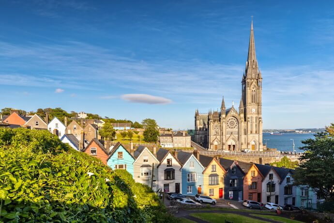 Colorful row houses with St. Colman’s Cathedral rising in the background, overlooking Cork Harbour on a sunny day.