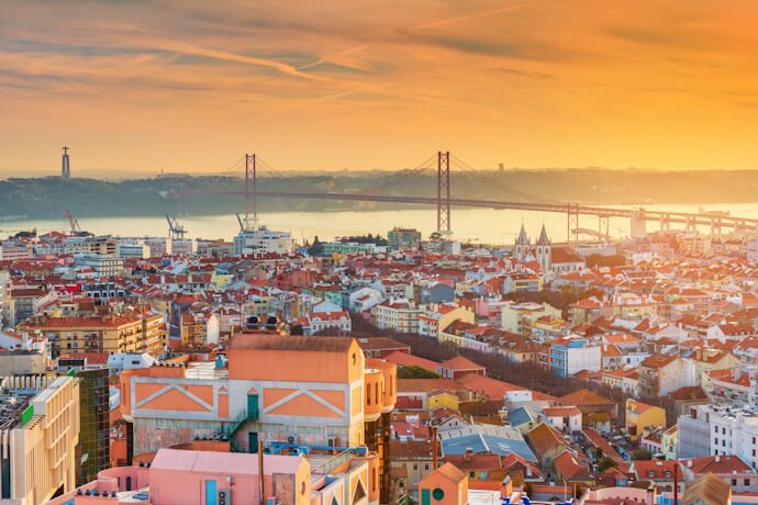 Panoramic sunset view over Lisbon rooftops and the Tagus River with the 25 de Abril Bridge in the background.