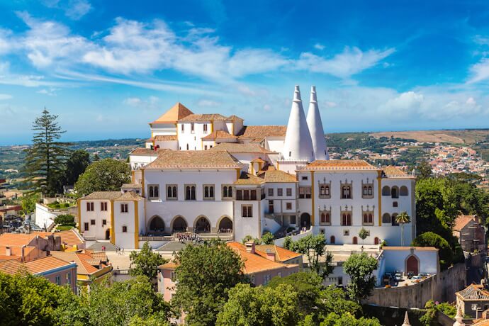 The white National Palace of Sintra with its twin chimneys overlooking the town’s red rooftops under a bright blue sky.