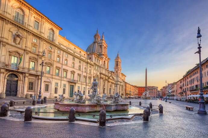 People relaxing at outdoor cafés around a colorful Roman piazza on a sunny day, enjoying coffee and lively atmosphere.