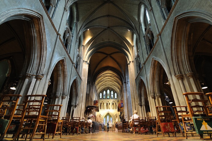 The interior of a historic Gothic church featuring high vaulted ceilings, stone arches, wooden chairs, and a softly illuminated altar at the far end.