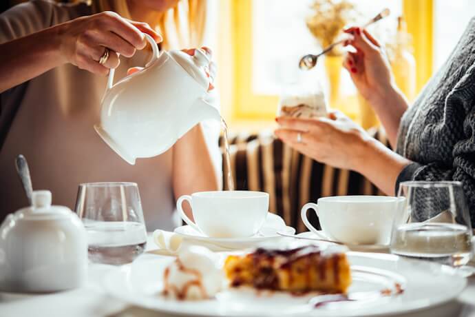 Two people enjoying afternoon tea, pouring from a teapot into cups with cake and pastries on the table.