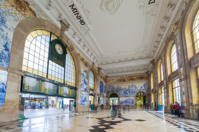Grand historic train station hall in Porto with ornate ceiling, large arched windows, and blue-and-white tile murals.