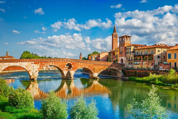 A picturesque view of Verona, Italy, featuring the ancient Ponte Pietra stone bridge arching over the Adige River with historic buildings and a bell tower in the background under a bright blue sky.