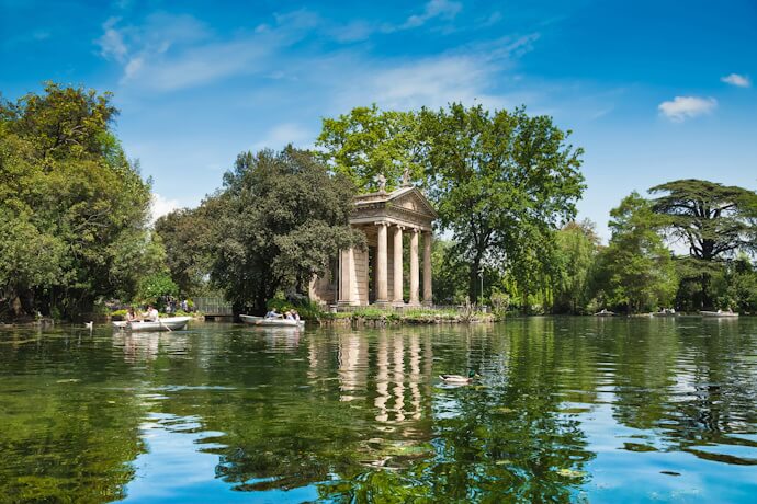 Temple of Aesculapius reflected in the lake at Villa Borghese Gardens in Rome, surrounded by lush trees and rowboats.
