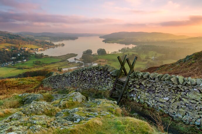 Sunset view over a Lake District lake and rolling fells, seen from a hillside beside a dry-stone wall and wooden stile.