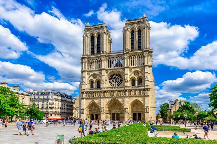 Front view of the Notre-Dame Cathedral in Paris on a sunny day, with a crowd of visitors gathered in the square and lush greenery in the foreground.”