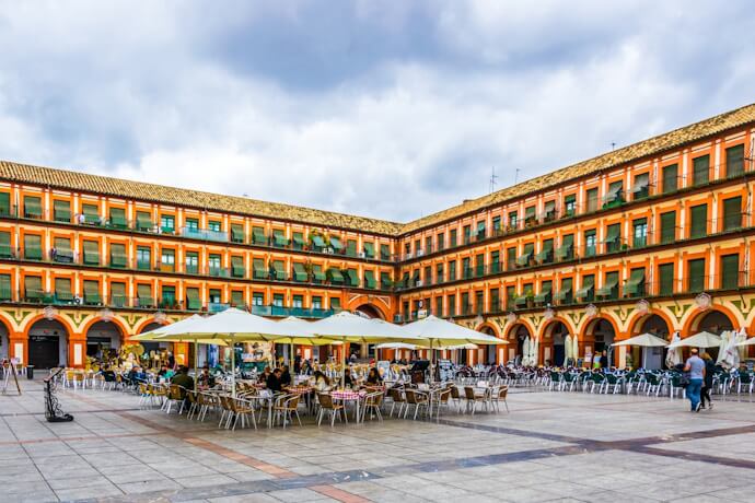 View of Plaza de la Corredera in Córdoba, Spain, with colorful arched façades, rows of balconies, and a central café area filled with people under large white umbrellas.