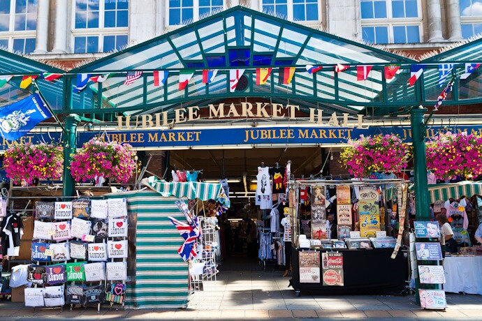 Colorful entrance to Jubilee Market Hall in London, decorated with international flags and hanging flower baskets, with stalls selling souvenirs, clothing, and food items.