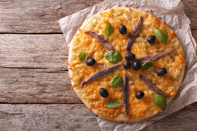 A round Pissaladière tart topped with caramelized onions, anchovies arranged in a star pattern, black olives, and fresh basil leaves, placed on parchment over rustic wooden boards.