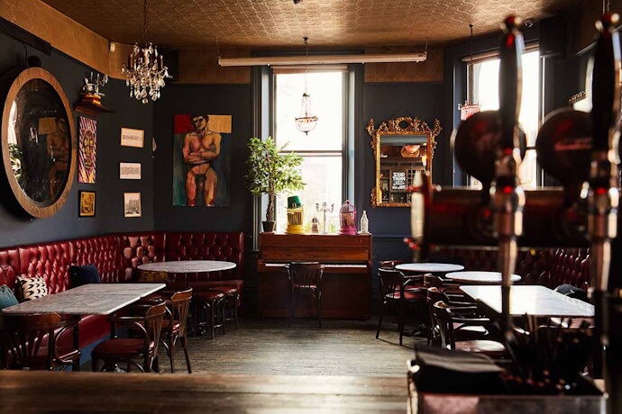 A cozy, dimly lit pub with vintage décor, dark walls, red leather seating, and eclectic artwork, including a portrait and a mirror reflecting warm light.