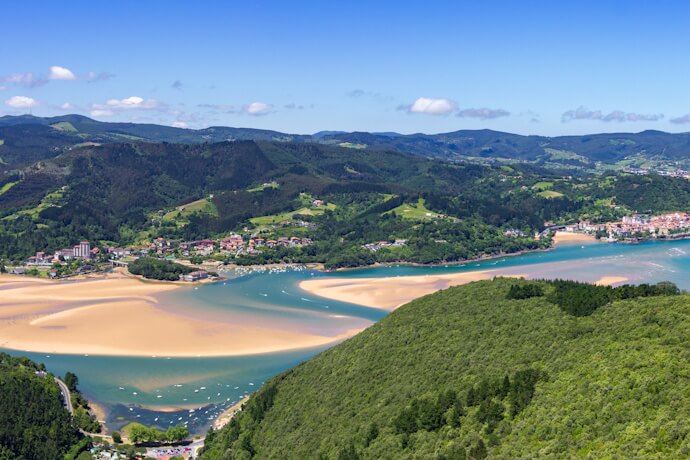 In the Urdaibai Biosphere Reserve, a broad estuary winds through green hills and sandy banks, forming a peaceful natural landscape dotted with small villages.