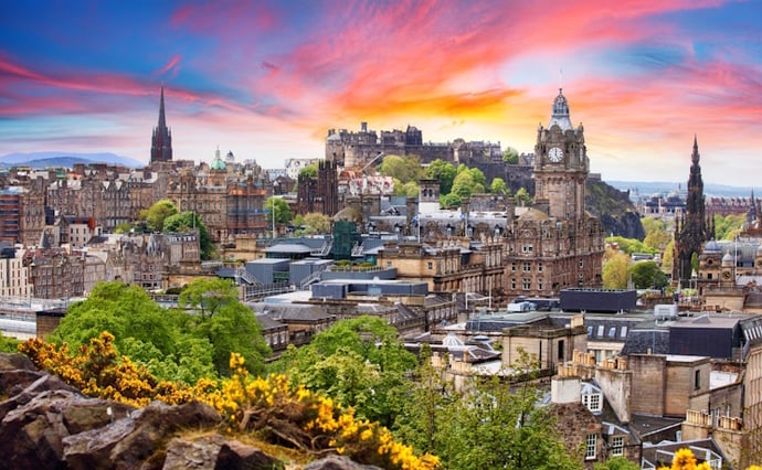 Sunset view of Edinburgh with historic buildings, the Balmoral clock tower, and Edinburgh Castle on a green hill.