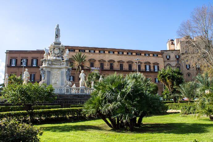 Palermo’s Royal Palace and fountain from a sunlit piazza, featuring lush palm trees and an ornate central monument
