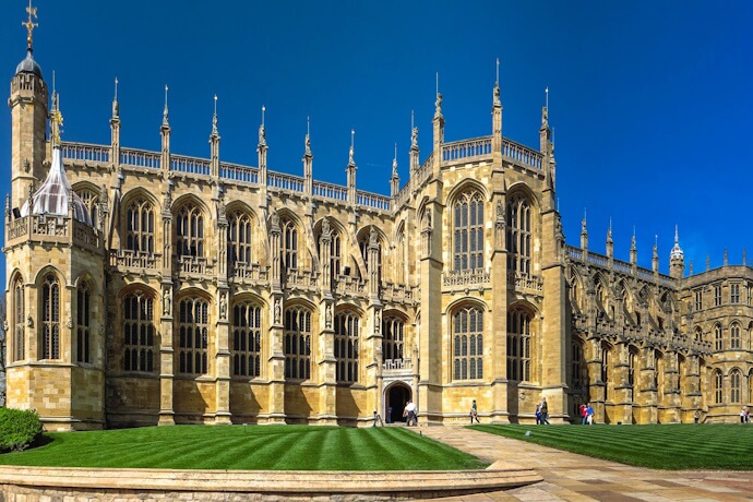 Exterior view of St George's Chapel at Windsor Castle in England, showcasing its grand Gothic architecture, tall windows, and manicured lawns under a clear blue sky.