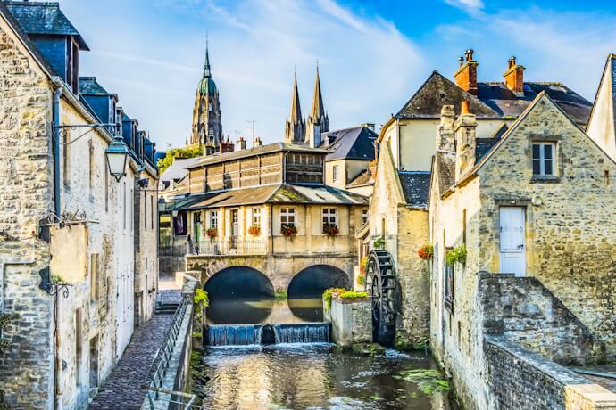 Charming stone houses and a quiet canal in Bayeux, with a historic watermill and cathedral towers rising above the medieval town.
