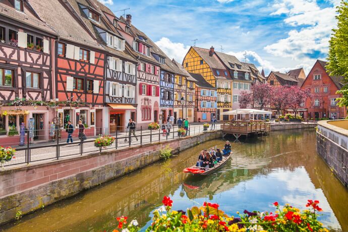 Colorful half-timbered houses lining a peaceful canal in Colmar, Alsace, with flower-filled balconies and a small boat gliding through the historic old town.