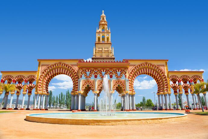 A grand entrance archway with Moorish-inspired architecture stands against a bright blue sky in Córdoba.