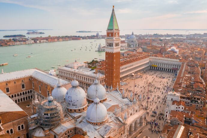Panoramic aerial view of Piazza San Marco in Venice, showcasing the domes of St. Mark’s Basilica, the towering Campanile, and the Grand Canal filled with boats in the distance.