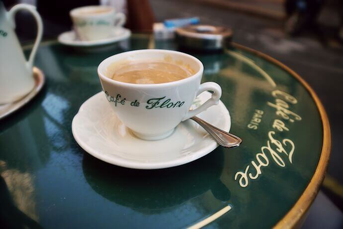 A close-up of a white porcelain cup filled with coffee, sitting on a matching saucer with a silver spoon from the Cafe de Flore in Paris.