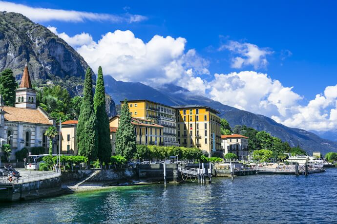 A picturesque lakeside town on Lake Como in Lombardy, Italy, featuring elegant yellow buildings, cypress trees, and a historic church tower, all set against a backdrop of dramatic alpine mountains and a bright blue sky.