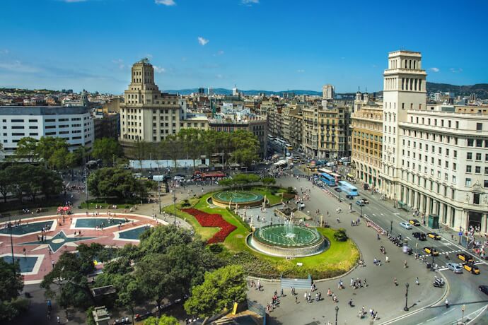 Wide view of Plaça de Catalunya in Barcelona, Spain, featuring fountains, green areas, and surrounding grand buildings, with busy pedestrian traffic and buses around the roundabout.
