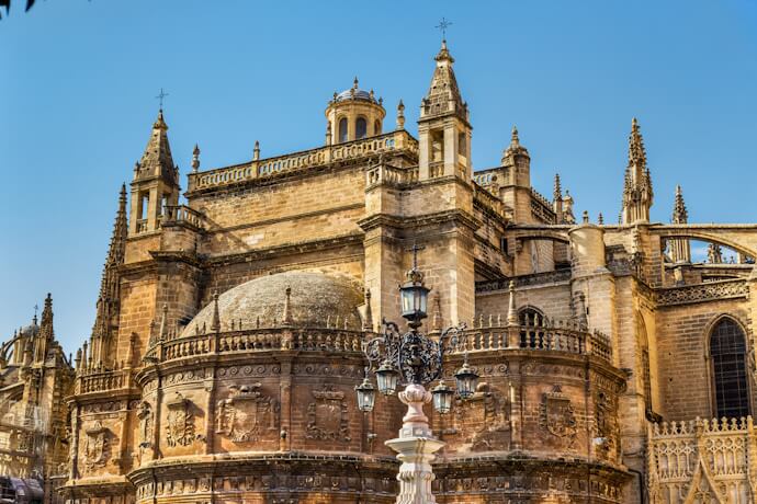Detailed view of Seville Cathedral’s ornate Gothic exterior, showcasing its stone carvings, domed roof, and spires under a clear blue sky.