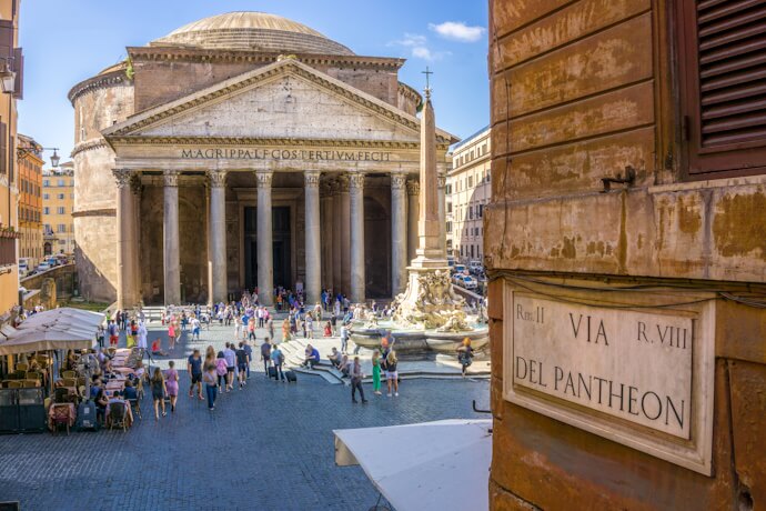 A lively scene in front of the Pantheon in Rome, showing crowds of visitors gathered near the ancient temple’s grand Corinthian columns. The inscription on the pediment is visible, and a tall fountain with an obelisk stands in the foreground.