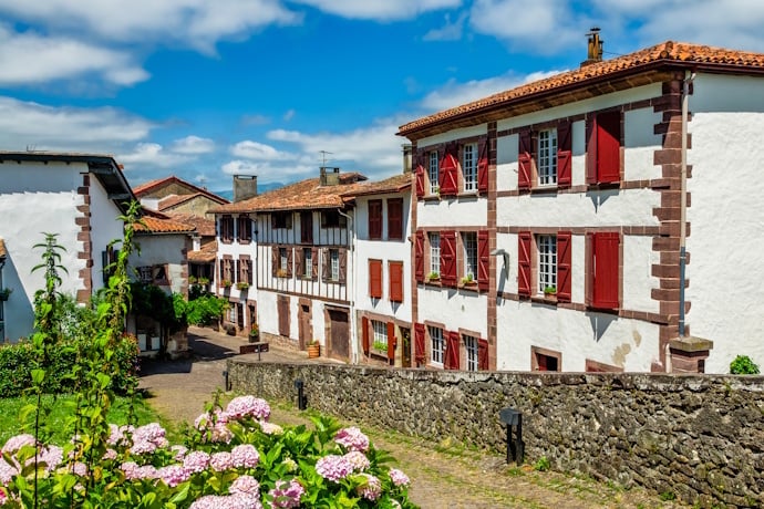 A scenic street in the Basque town of Saint-Jean-Pied-de-Port, France, lined with traditional whitewashed houses with red shutters and cobblestone paths surrounded by blooming hydrangeas.