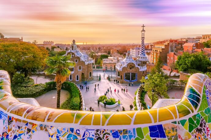 View over Park Güell in Barcelona at sunset, featuring Gaudí’s colorful mosaic terrace, whimsical architectural structures, and panoramic views of the city with a warm, glowing sky.