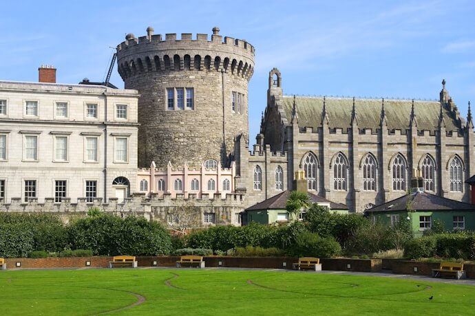 The medieval round tower and Gothic chapel of Dublin Castle surrounded by manicured lawns and historic buildings under a bright blue sky.