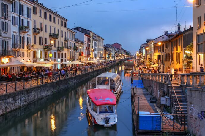 Evening view of the lively Navigli district in Milan, Italy, with a canal lined by vibrant buildings and outdoor restaurants full of people.