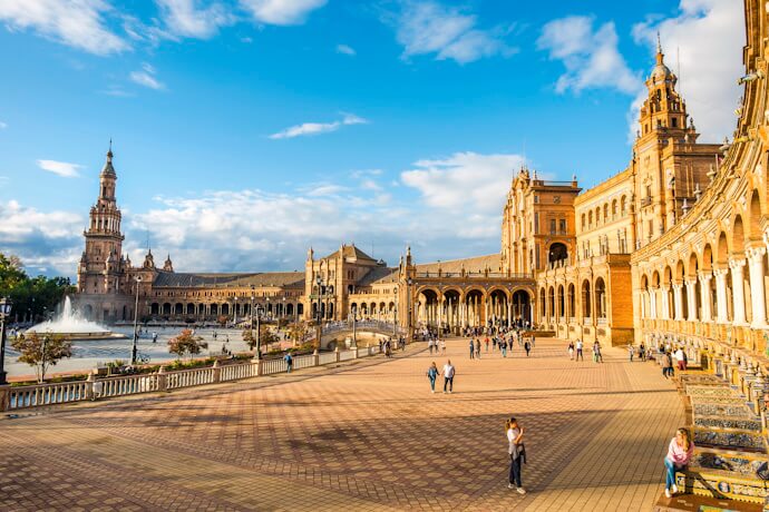 Elegant semicircular square of Plaza de España in Seville, Spain, with Renaissance-style architecture, towers, and arcades, alongside a canal and central fountain with visitors strolling.
