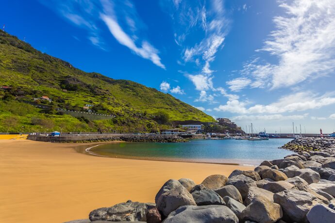 Calm sandy beach in Machico Bay, Madeira, with clear blue water, green hills in the background.