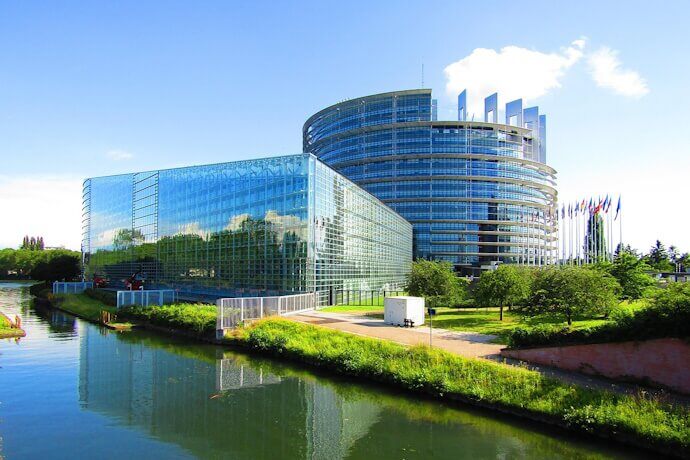 European Parliament building in Strasbourg, a modern glass and steel structure with a curved façade reflecting the sky and surrounding greenery. The striking circular design symbolizes unity, while the European flags flutter in front.