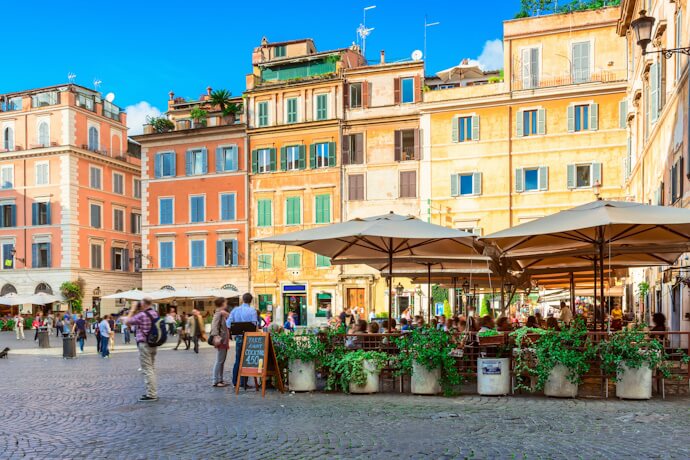 A vibrant piazza in the Trastevere neighborhood of Rome, featuring colorful historic buildings with shuttered windows and a lively outdoor café.
