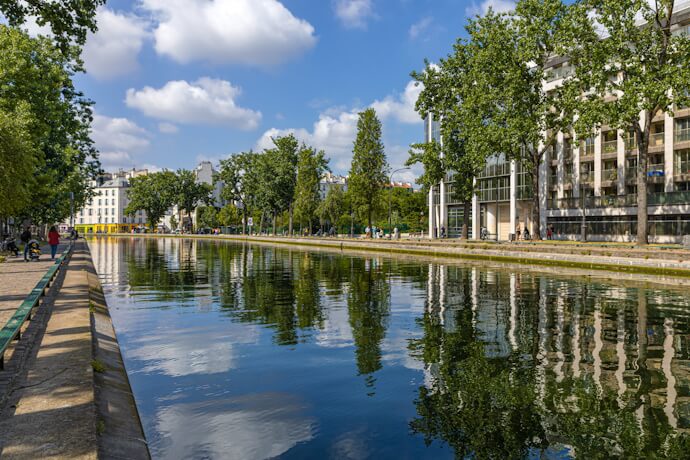 Tranquil view of the Canal Saint-Martin in Paris with reflections of trees and buildings in the calm water, bordered by walkways and green spaces.