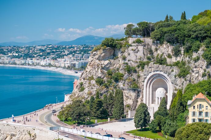 A sweeping view of the coast of Nice from Castle Hill, showing the azure Mediterranean, a road curving along the cliffs, and the grand Monument aux Morts carved into the rock face.