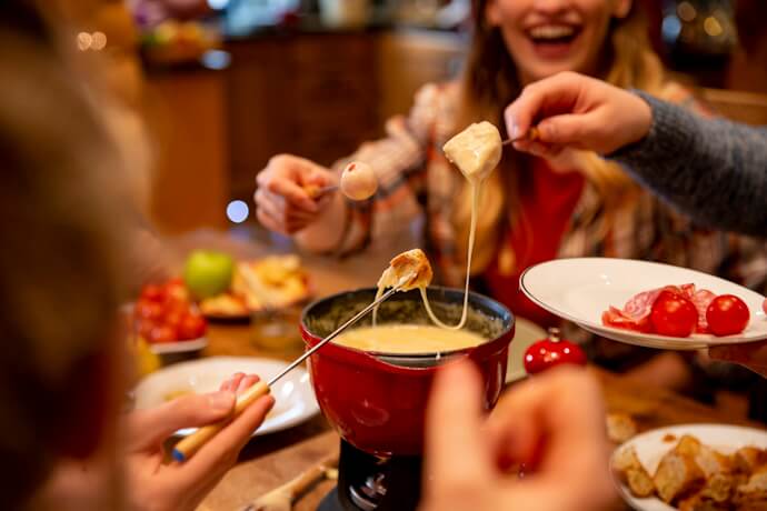 Friends sharing a pot of melted cheese fondue with bread, tomatoes, and charcuterie in a cozy setting.