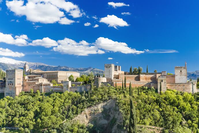 Panoramic view of the Alhambra palace complex in Granada set against green hills and snow-capped mountains under a blue sky.