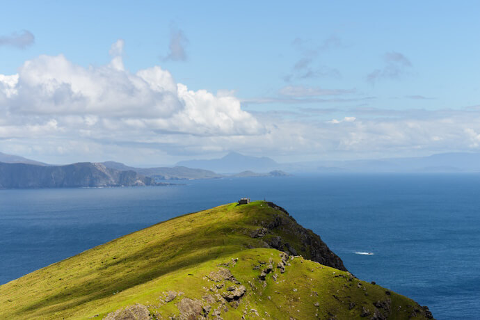 A grassy headland extending into the deep blue ocean with distant mountains on the horizon.