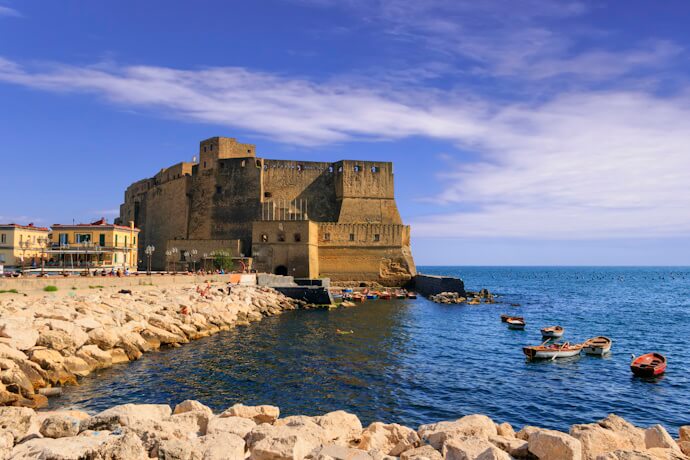 View of Castel dell'Ovo on the seafront in Naples, Italy, with small boats floating in the blue waters and people relaxing on the rocky shoreline.
