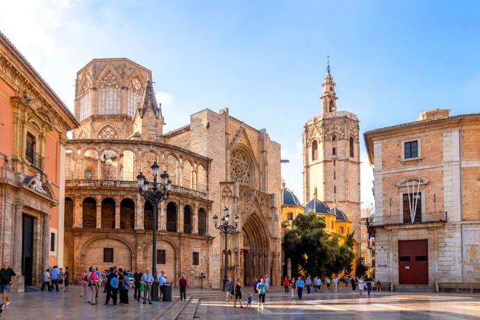 Historic square in Valencia, Spain, known as Plaza de la Virgen, with the Valencia Cathedral and Basilica, Gothic and Baroque architecture, and groups of tourists exploring the area.