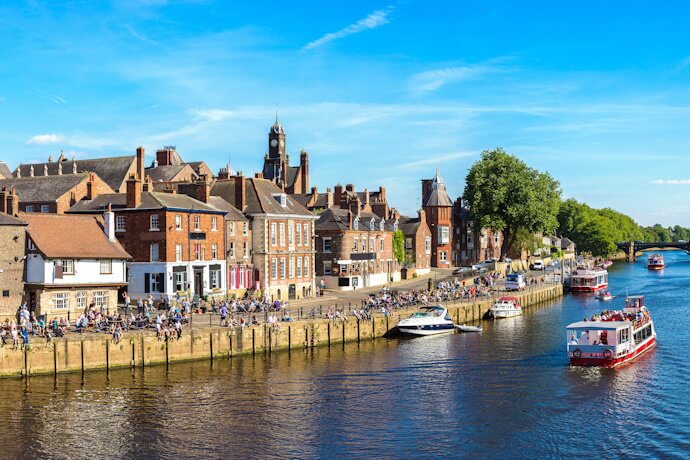 Scenic view of the River Ouse in York, with historic riverside buildings, boats cruising on the water, and people enjoying the sunny day along the promenade.