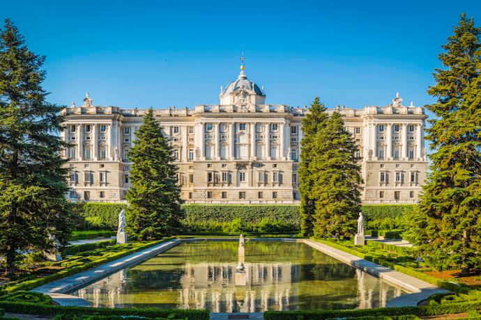 Front view of the grand Royal Palace in Madrid with manicured gardens and a reflecting pool in the foreground.