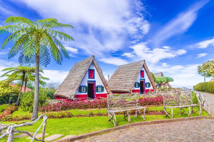 Traditional triangular thatched-roof houses in Santana, Madeira, surrounded by lush gardens, flowers, and cobblestone paths.