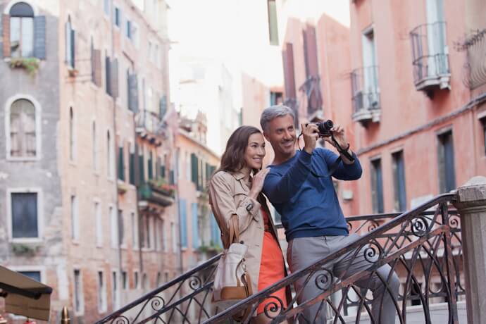 Smiling couple enjoying a moment on a quaint Venetian bridge, surrounded by narrow canals and colorful historic buildings with shuttered windows.
